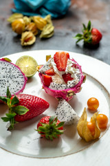 on a white, wooden plate, lie the fresh fruit of strawberries, among them a beautiful, pink dragon fruit filled with whipped cream and strawberries