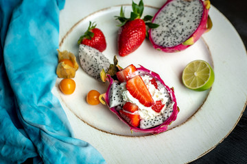 on a white, wooden plate, lie the fresh fruit of strawberries, among them a beautiful, pink dragon fruit filled with whipped cream and strawberries