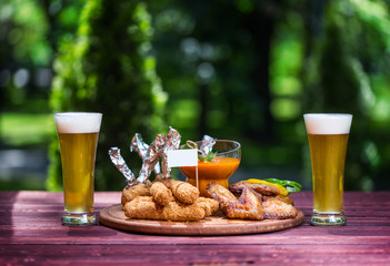 Chicken and Cheese Set for beer and two glasses of beer. Chicken wings, cheese sticks and cheese balls with bright vegetable sauce. On the cutting board, wooden and green summer background