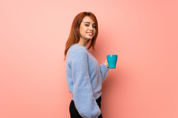 Young redhead woman over pink background holding a hot cup of coffee