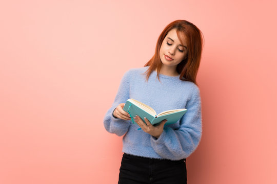 Young redhead woman over pink background holding a book and enjoying reading