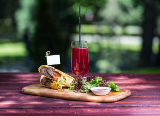 Healthy shawarma sandwich with beef, green salad, sauce and lemonade. On the cutting wooden board, and green summer background.