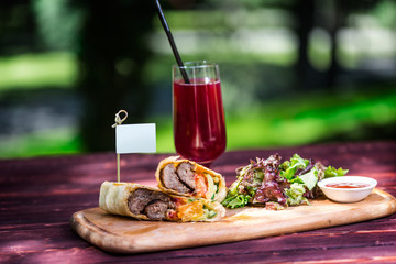 Healthy shawarma sandwich with beef, green salad, sauce and lemonade. On the cutting wooden board, and green summer background.