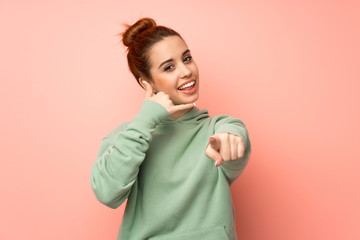 Young redhead woman with sweatshirt making phone gesture and pointing front