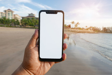 Close up of hand holding phone with white screen by the beach. Smartphone with mockup on background beach and hotel or resort.
