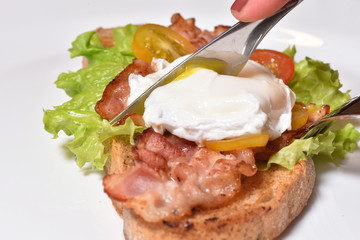 Female hands cut a fried egg with a knife and a beautiful sandwich with fried bacon and cherry tomatoes. Unhealthy food for breakfast. CLose-up