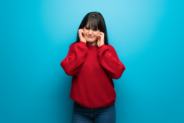 Woman with red sweater over blue wall frustrated and covering ears with hands