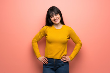 Woman with yellow sweater over pink wall posing with arms at hip and smiling