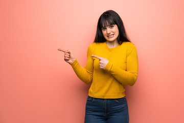 Woman with yellow sweater over pink wall frightened and pointing to the side