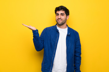 Man with blue jacket over yellow wall holding copyspace imaginary on the palm to insert an ad