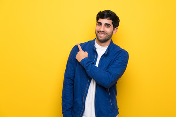 Man with blue jacket over yellow wall pointing to the side to present a product