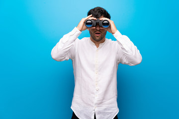 Young man over isolated blue wall and looking in the distance with binoculars