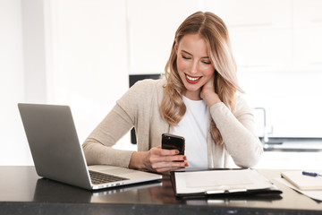 Blonde woman posing sitting indoors at home using laptop computer and mobile phone.