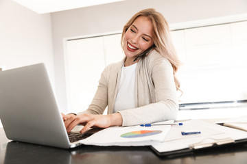 Beautiful blonde woman posing sitting indoors at home using laptop computer talking by mobile phone.