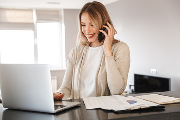 Blonde woman posing sitting indoors at home using laptop computer talking by mobile phone.
