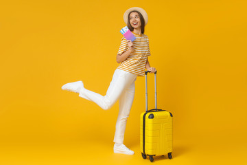 Funny girl jumping, holding suitcase and passport with flight tickets, isolated on yellow background