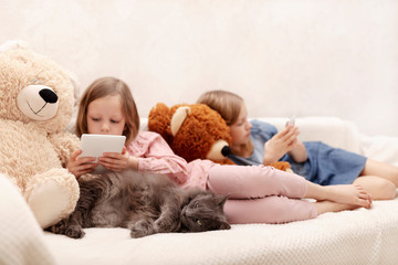 Two little girls (sisters 7 and 8 years old) are watching an e-book and a smartphone at home on the sofa with their cat.
