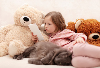 Two little girls (sisters 7 and 8 years old) are watching an e-book and a smartphone at home on the sofa with their cat.