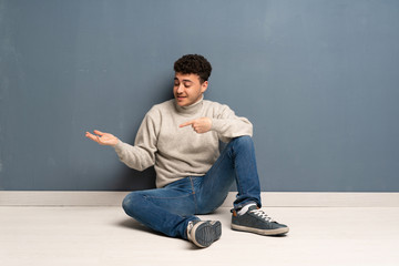 Young man sitting on the floor holding copyspace imaginary on the palm to insert an ad