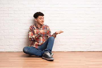 Young man sitting on the floor holding copyspace imaginary on the palm to insert an ad