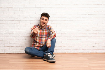 Fototapeta na wymiar Young man sitting on the floor inviting to come with hand. Happy that you came