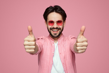 Portrait of trendy young man in pink stylish shirt and sunglasses showing thumb up over pink...