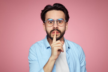 Fashionable hipster guy demands keeps silence, shows hush gesture, looks with stunned expression at camera, afraids of gossiping, isolated over pink background. Mute man