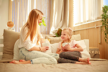 Mother and daughter with cup of tea at home talking on bed, happy family lifestyle