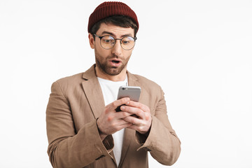 Photo of excited man 30s wearing hipster hat smiling while holding and using smartphone