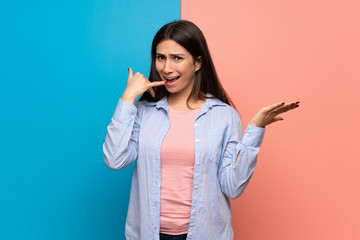 Young woman over pink and blue wall surprised and making phone gesture