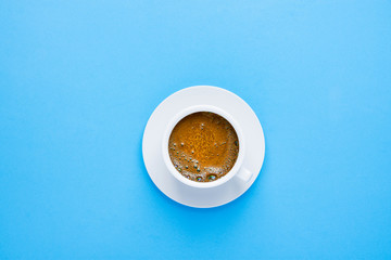 One white cup of freshly brewed black coffee with tan-colored crema foam on saucer on light blue background. Top view. Morning breakfast energy caffeine addiction.