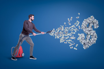 Side view of man in casual clothes hoovering up dollars and big dollar sign with vacuum cleaner.
