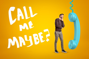 Young man in casual clothes standing in half-turn, one hand on chin, near big cyan phone receiver...