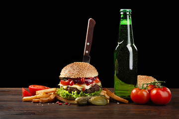 Homemade hamburger with french fries and bottle of beer on wooden table. In the burger stuck a knife. Fastfood on dark background