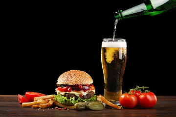 Homemade hamburger with french fries and bottle of beer pouring into a glass. Fastfood on dark background on wooden table