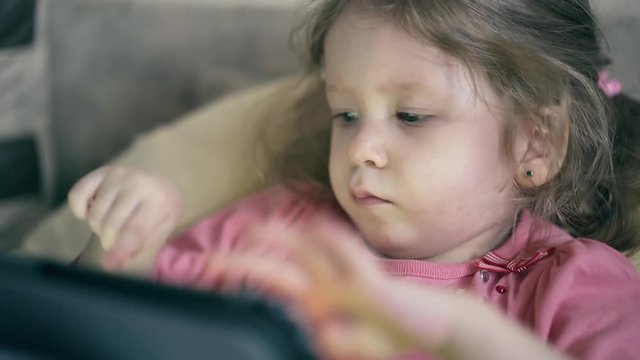 CU: portrait of pretty little girl, child who lies on the sofa under the blanket and plays on a tablet computer, sometimes reflects.
