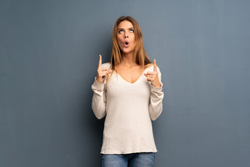 Blonde woman over grey background pointing up and surprised