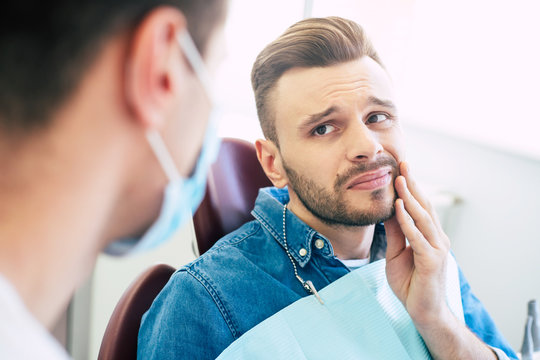 Symptoms of gums pain. A man with a worried face is holding his hand on his cheek  because of irritating pain in front of a dentist who is going to give a patient a treatment.