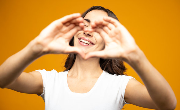 Center of emotions. Pretty girl next to the yellow background is showing a heart with her hands and smiling sincerely right in the center of this heart. - Powered by Adobe