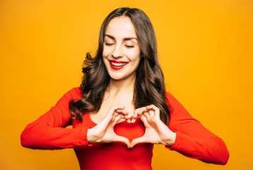 To my boyfriend. Fabulous girl is showing a heart shape with her hands in front of orange background wearing vibrant red sweater and simple makeup.