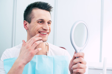 Nice and neat. A photo of pleased man in a dentist cabinet with a mirror in his hand which hi is using for observing the final result of the treatment and he looks absolutely satisfied.
