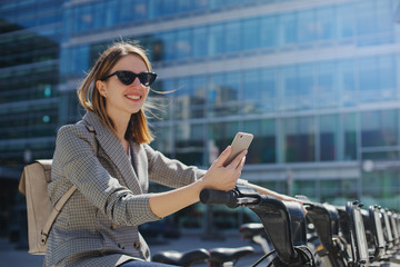 Happy smiling woman using bicycle rent mobile app outdoors, Portrait of female manager browsing smartphone while seating on bike