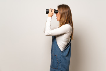 Beautiful woman over isolated white wall and looking in the distance with binoculars