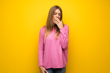 Woman with pink sweater over yellow wall yawning and covering wide open mouth with hand