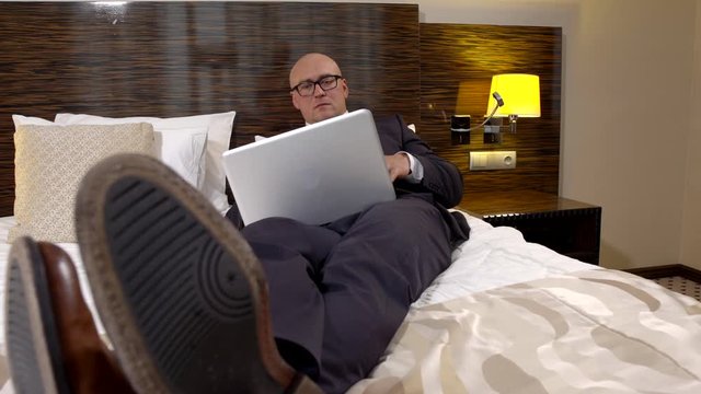 business bald man with glasses and a stylish business suit, lying on the bed in the hotel room and looking at the laptop, next to him is the phone. Shoes men close-up.