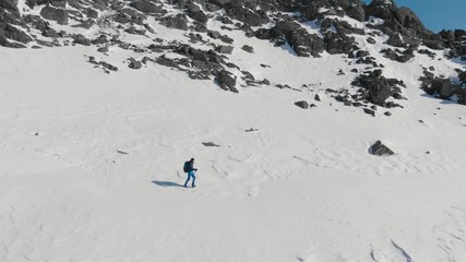 Aerial view of young man hiking climbing snowy mountain at beautiful winter day. Happy active lifestyle video. Male mountaineer with trekking poles and a backpack walking on mountain ridge.