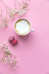 Flatlay pink coral background, the cup of cappuccino coffee and sweets macaroons, spring white flowers
