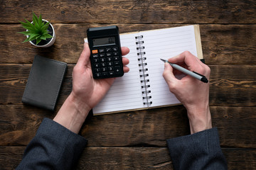 Business man is counting money on a calculator and is writing a sum in a notebook with a blank...