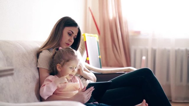 Travelling, 4k: Young attractive mother and sweet daughter are sitting on the couch and training using a tablet computer. The daughter thinks. Smiling.