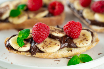 Front view of toast bread with topping - chocolate paste, bananas, raspberries and mint on white background. Tasty sweet summer breakfast with toasts close-up